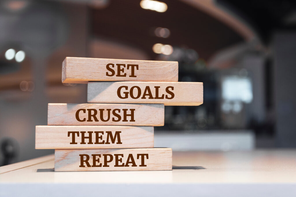 Stacked wooden blocks with the motivational phrase 'Set Goals, Crush Them, Repeat' printed in bold letters, placed on a table with a blurred background.